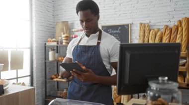 Fırında çalışan genç adam, etrafında hamur işleri, raflar ve modern bir yazar kasayla çevrili bir tablet kullanıyor.