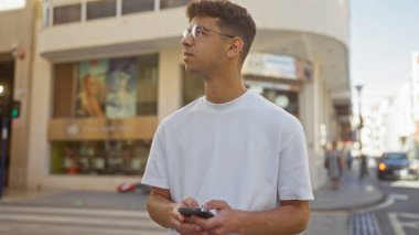 Handsome young hispanic man holding a smartphone with a contemplative expression on an urban street during daytime. clipart