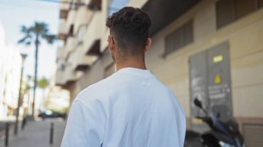 Beyaz gömlekli, genç, yakışıklı, İspanyol bir adam arka planda palmiye ağaçları ve binaları olan bir şehir sokağında duruyor..
