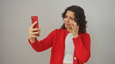 Kıvırcık esmer saçlı, kırmızı kazak giyen, komik bir yüz ifadesi takınırken kırmızı akıllı telefonuyla selfie çeken, beyaz bir arka planda izole edilmiş İspanyol bir kadın..