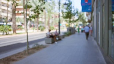 Ağaçlar ve binalarla çevrili güneşli bir kaldırımda yürüyen ve oturan insanların olduğu bulanık bir açık hava sahnesi..