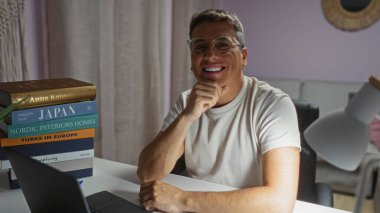 Young man smiling in a living room interior with a stack of books on a desk clipart