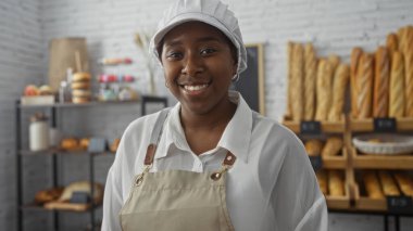 Güzel, Afro-Amerikan kadın pastanesi. Kapalı mekanda, ekmek sergisi önünde önlük ve şapka takarak rahatça poz veriyor.