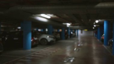 Bulanık yeraltı otoparkı, mavi sütunlar ve loş ışıklı, bulanık ve odak dışı arka planda park etmiş arabalar..