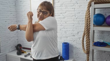 Orta yaşlı İspanyol bir kadın spor salonunda arka planda ekipmanlarıyla kolunu uzatıyor. Gözlük takıyor ve beyaz tişört giyiyor..