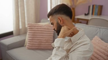 Sakallı, rahat oturma odasında oturmuş, evde dinlenirken boynunu kaşıyan genç İspanyol bir adam.