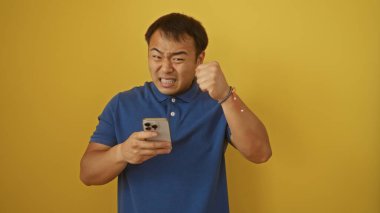 Mavi tişörtlü genç Çinli adam telefonuna bakarken heyecanlanıyor, sarı arka planda duruyor..