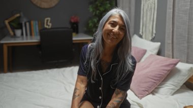 Beyaz saçlı bir kadın, yatak odasında rahat bir yatakta oturuyor. Yastıkları ve arka planda bir masası var.