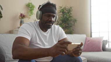 Yakışıklı, sakallı, oturma odasında dinlenen, kulaklık takan ve rahat bir dairede akıllı telefon kullanan Afro-Amerikan bir adam..