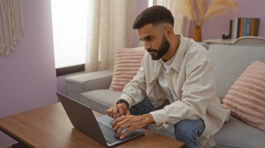 Evde dizüstü bilgisayarla çalışan sakallı genç İspanyol bir adam, rahat bir oturma odasında pembe yastıklı ve hafif ışıklandırmalı bir koltukta oturuyor..
