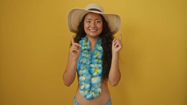 Çiçekli çiçek kolyesi takan güzel Asyalı bir kadın ve sarı izole bir arka plana gülümserken hasır bir şapka yukarı bakıyor.