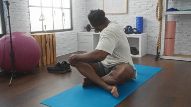 Afrika kökenli Amerikalı bir adam modern bir spor salonunda yoga yapıyor. Yanında spor topu, ayakkabı ve ağırlık gibi ekipmanlar var. Yoga minderinde, rahat ve iyi aydınlatılmış bir odada oturuyor..