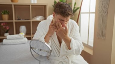 Sağlık kaplıcasında genç bir adam yüzünde sivilce patlatırken yüz temizleme seansı için beyaz bornoz giyiyor..