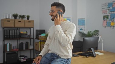 Ofisinde kitaplıklar ve ofis malzemeleriyle otururken telefonda eğlenen yakışıklı İspanyol bir adam..