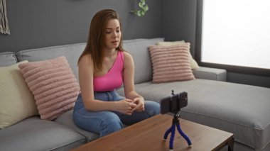 Pembe bluzlu genç kadın, rahat oturma odasında, gri kanepesi ve akıllı telefonunda pembe yastıkları olan bir video kaydediyor.