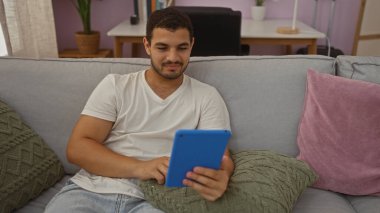 Genç İspanyol bir adam oturma odasında rahat bir kanepede otururken tablet üzerinde geziniyor.