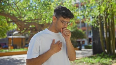 Arka planda ağaçlar ve şehir binaları olan bir şehir parkında öksüren İspanyol bir adam.