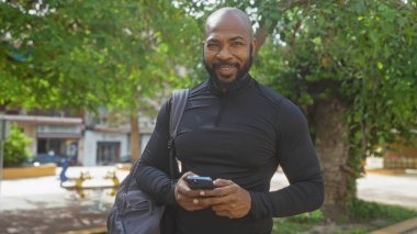 Sakallı yakışıklı kel adam şehir parkında elinde akıllı bir telefonla dikiliyor.