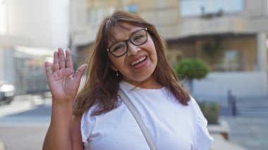 Neşeli orta yaşlı İspanyol bir kadın şehir sokaklarında beyaz bir gömlek ve gözlük takıyor ve modern bir şehir geçmişine sahip..