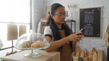 Genç Asyalı kadın fırında akıllı telefon kullanıyor, arka planda hamur işleri ve ekmek var.