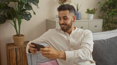 Yakışıklı, genç, İspanyol bir adam rahat oturma odasında kanepede otururken gülümsüyor, ev içinde sıcak bir şekilde aydınlatılmış akıllı telefonuyla meşgul oluyor..