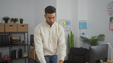 Kapalı bir ofiste masa başında çalışan genç, İspanyol bir adam. Organize rafları ve ofis bitkileri olan modern bir ofis..