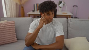 Modern bir oturma odasında, şakağına parmağını koymuş, kanepede oturan yakışıklı İspanyol bir adam.