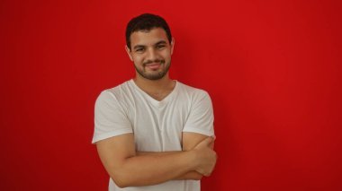 Yakışıklı, genç, İspanyol bir adam, canlı kırmızı bir arka plana karşı kollarını kavuşturarak güvenle gülümsüyor.