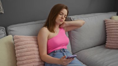 Pembe bluzlu genç bir kadın oturma odasındaki gri bir koltukta oturuyor. Elinde akıllı bir telefonla boynuna acı içinde dokunuyor.
