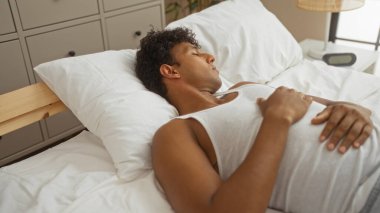 Rahat bir yatak odasında huzur içinde uyuyan genç bir adam, rahat beyaz bir yatakta uzanıyor rahat bir yüz ifadesi ve sakin bir atmosferi var..