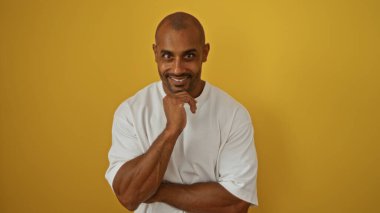 Yakışıklı, Afro-Amerikalı bir adam izole edilmiş sarı bir arka planda gülümsüyor, beyaz bir gömlek giyiyor ve çenesinin altında eli ile güven içinde poz veriyor..