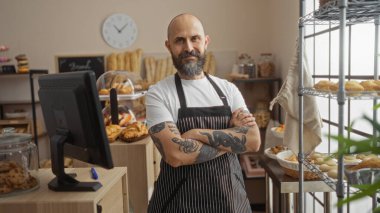 Sakallı ve dövmeli İspanyol bir adam, çapraz kollu bir fırında güvenle duruyor, raflarda çeşitli pastalar ve tezgahlarla çevrili, kapalı bir dükkan ortamını ön plana çıkarıyor..