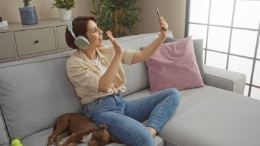Oturma odasında evcil köpeği yanında kanepede yatarken video sohbeti yapan kulaklıklı beyaz bir kadın..