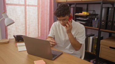 Ofis ortamında genç, İspanyol bir adam telefonuyla konuşurken kredi kartına bakar ve ofis malzemeleriyle çevrili bir dizüstü bilgisayarla çalışır..