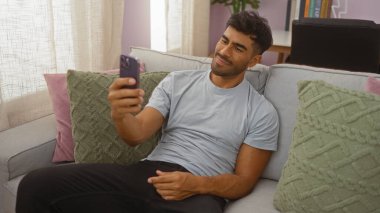 Yakışıklı genç İspanyol adam oturma odasında dinleniyor, akıllı telefonuyla selfie çekiyor, yeşil yastıklarla süslenmiş bir kanepede rahatça oturuyor..