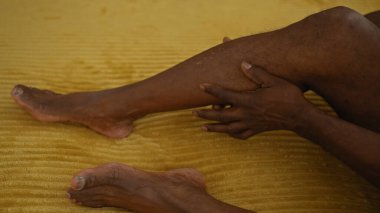 Afro-Amerikalı bir adam evdeki sarı bir yatakta yatarken bacağına dokunuyor..