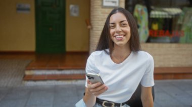 Şehir ortamında elinde akıllı bir telefonla gülümseyen genç bir kadın canlı bir şehir ortamını gözler önüne seriyor.