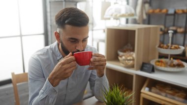 Samimi bir fırında kahvesini yudumlayan sakallı genç İspanyol adam içeride oturmuş hamur işleri ve ekmekle dolu rafların yanında.