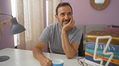 Yakışıklı, olgun İspanyol bir adam, oturma odasında düşünceli bir şekilde oturuyor, elinde kitaplarla bir fincan kahve ve masanın üzerinde bir lamba tutarken dikkatlice yukarı bakıyor.