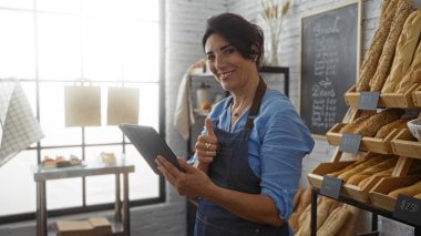 Fırında elinde tablet olan ve gülümseyen, çeşitli ekmek ve hamur işlerini sergileyen bir kadın.