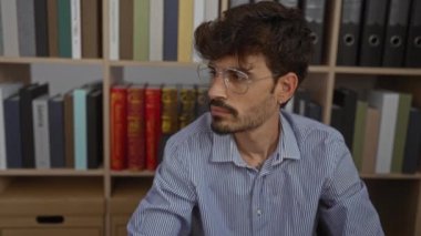 Ofiste sakallı, gözlüklü ve mavi çizgili tişörtlü bir genç var. Arka planda kitap ve klasörler var..