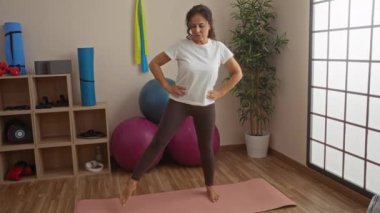 Kadın spor salonunda yoga minderi ve ekipmanlarıyla egzersiz yapıyor. Etrafı spor malzemeleri ve bitkilerle çevrili. Huzurlu bir ortamda..