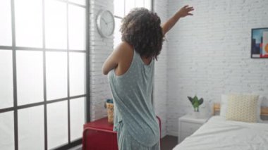 Yatak odasında geniş pencerenin ve beyaz tuğlalı duvarın yanında uzanan, modern, rahat bir evin içinde mavi pijama giyen bir kadın.