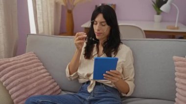 Modern bir oturma odasında rahat bir koltukta oturan genç, güzel İspanyol kadın mavi tabletini okuyor ve sakin bir ev ortamında rahat yastıklarla gözlüklerini ayarlıyor..