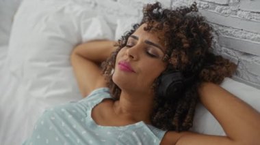 Yatak odasındaki kulaklıklarla yatakta dinlenen kadın içeride müzik dinlemekten zevk alıyor. Mutluluk ve sükuneti gösteriyor.