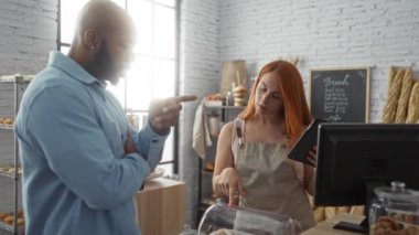 Fırıncı, arka planda ekmek ürünleri sergilenen tablet kullanarak müşteriye yardımcı olurken, kadın ve erkek bir fırında etkileşime giriyorlar.