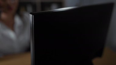 Ofiste telefonla konuşan esmer kadın arka planda kitap rafı olan kapalı alandaki bilgisayar ekranına bakıyor.