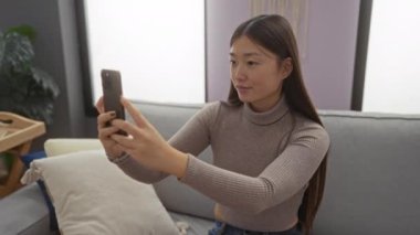 Oturma odasında akıllı telefon kullanarak selfie çeken bir kadın evinde modern dekorasyon ve doğal ışıkla çevrili bir anı yakalıyor.