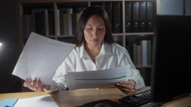 Ofiste çalışan genç İspanyol kadın lambanın altında belgeler okuyor, odaklanmış ve stresli görünüyor.