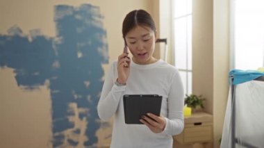 Yeni dekore edilmiş bir evde elinde tabletle telefonla konuşan bir kadın, iç tasarım sürecini ve teknoloji meraklısı çoklu görevleri sergiliyor..
